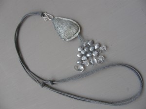 Halsband i fiskskinn (fettgarvat gös-skinn) och strandsten som tyngd inuti, med fejk-mocka-band, och "tofs" av vaxad lintråd, sötvattenspärlor och bergkristall. "Kroken" är av finsk sterling silver.